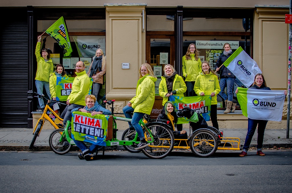 Das Büroteam des BUND Dresden steht in neonfarbenen BUND-Jacken, einem grünen lastenrad und BUND-fahnen vor dem Büro auf der Kamenzer Straße.